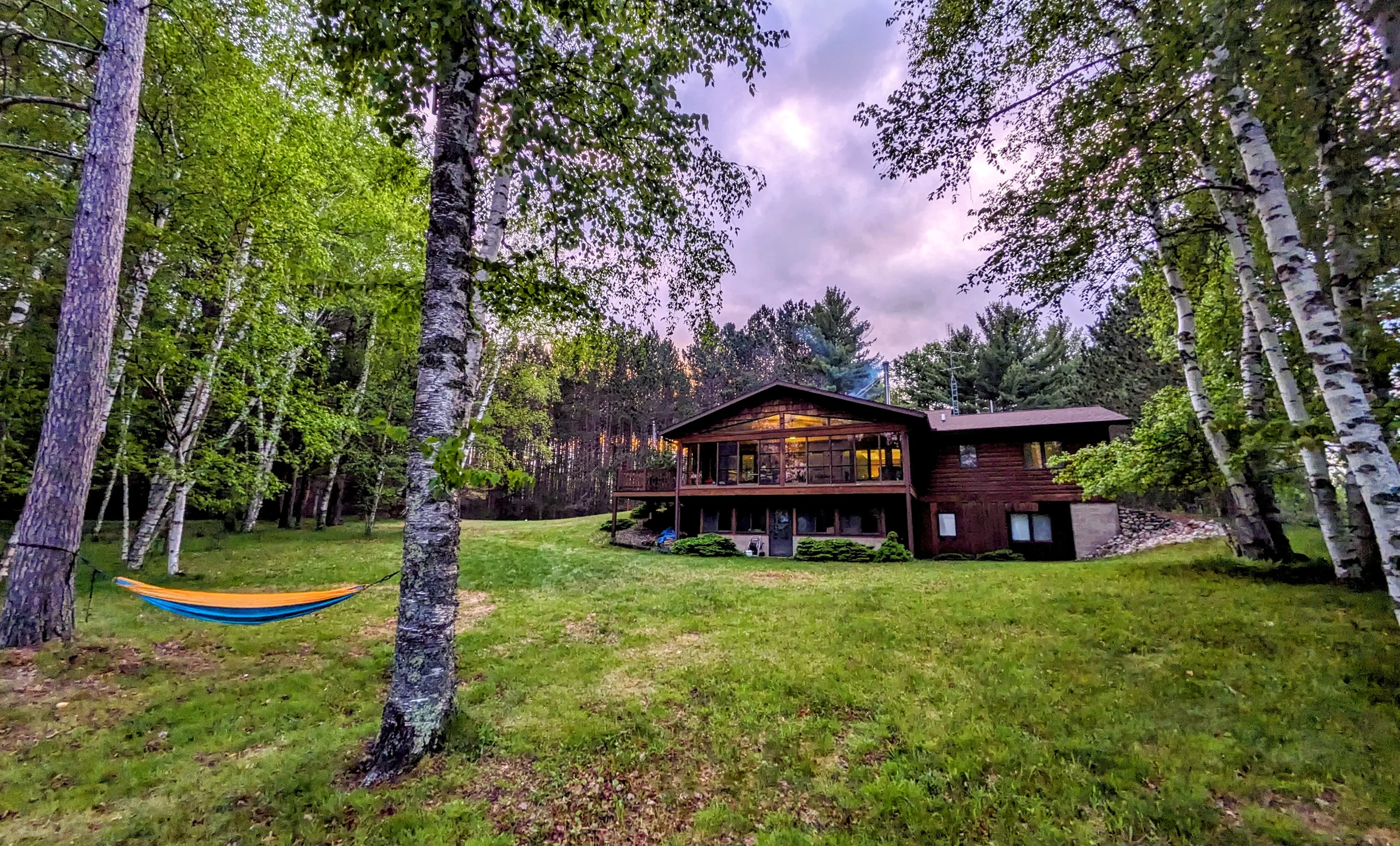 Snowden Lake home glowing at evening, framed by birch trees with a hammock on the lawn